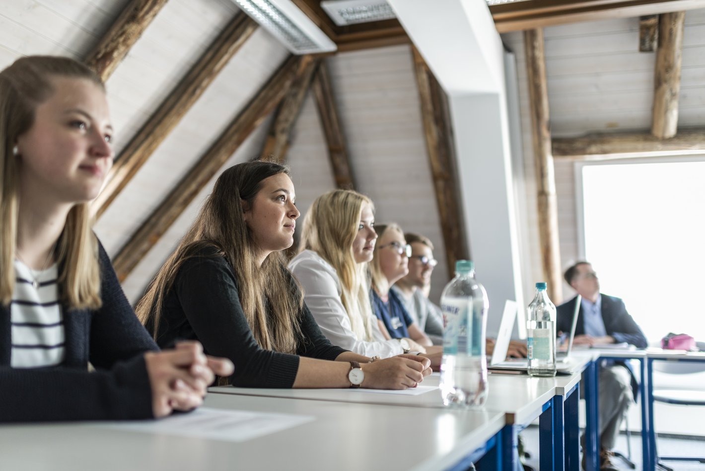 Gruppe von Studierenden in einem Seminarraum, eine Studentin ist im Fokus und hört aufmerksam etwas zu.