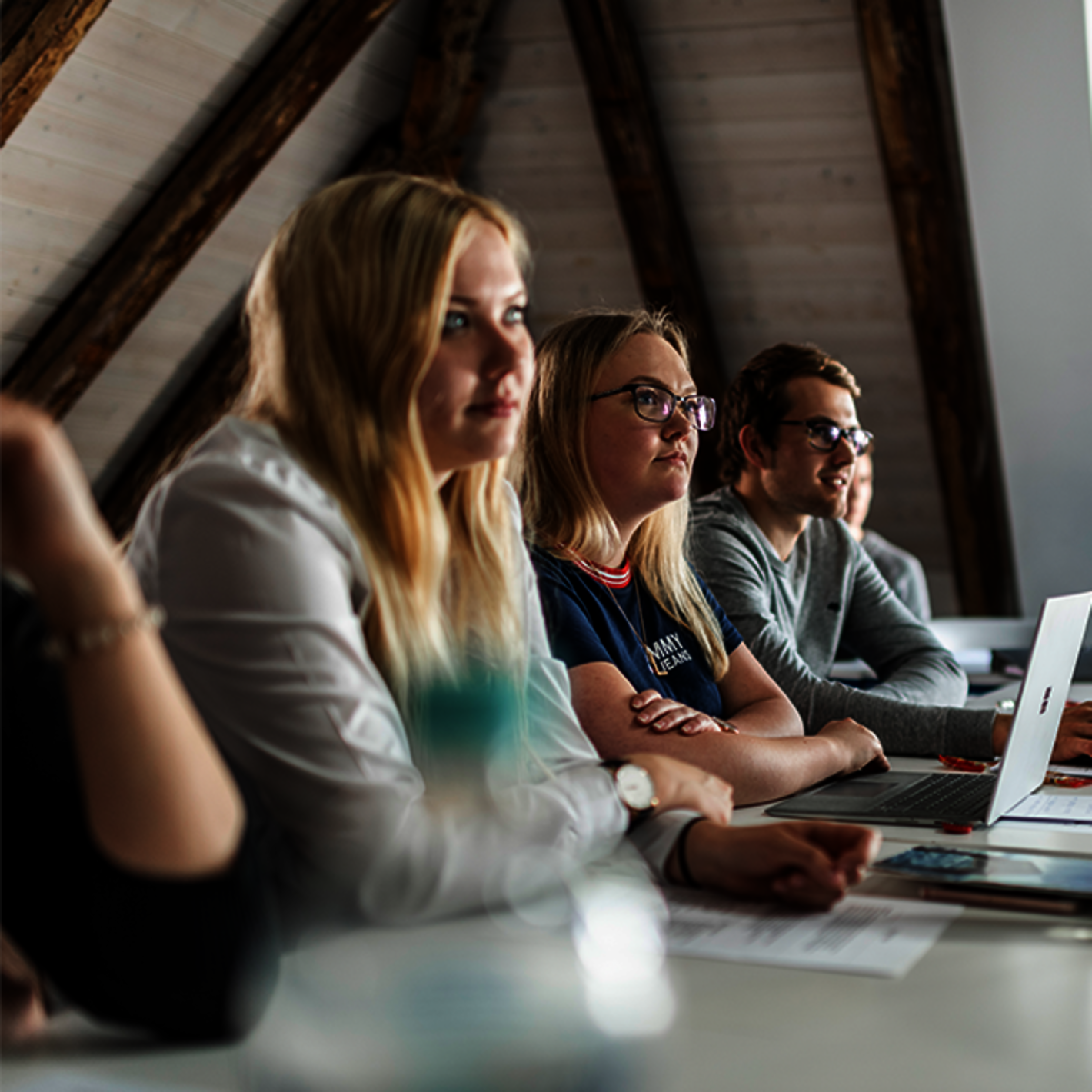 Studierende sitzen an einem Tisch und schauen zu einem Lehrenden
